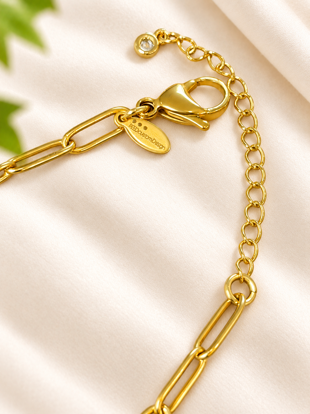 Close-up of a gold chain necklace with a lobster clasp, paperclip links, and a small oval brand tag on a soft cream fabric background.