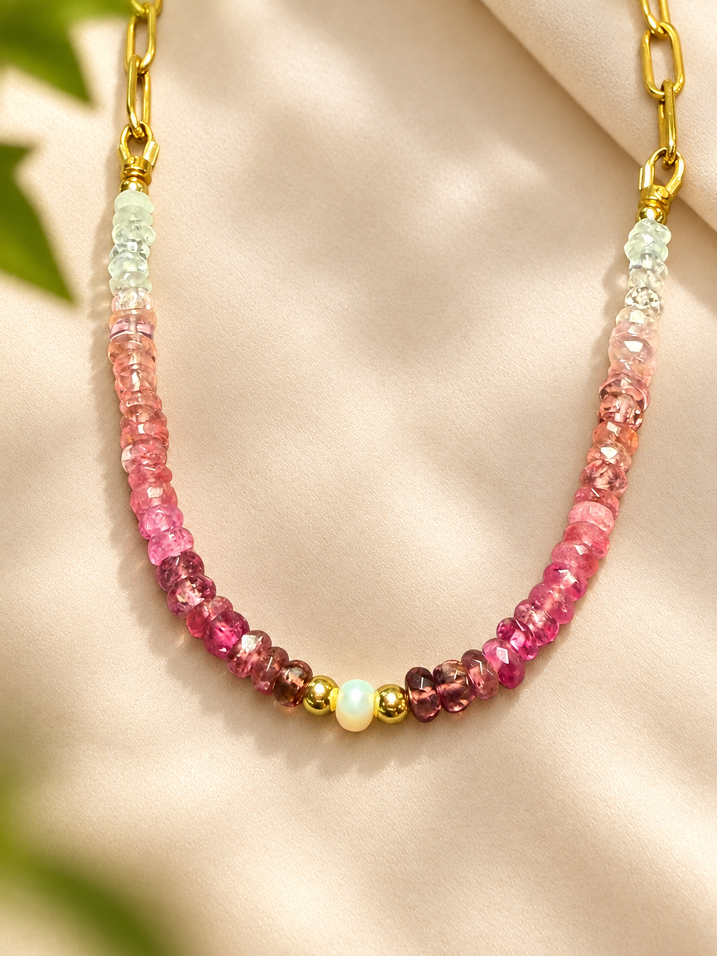 A beaded necklace featuring a gradient of pink and purple gemstone beads, a central cream-colored bead, and gold spacer beads, with gold chain links at the top, displayed on a beige satin fabric background.