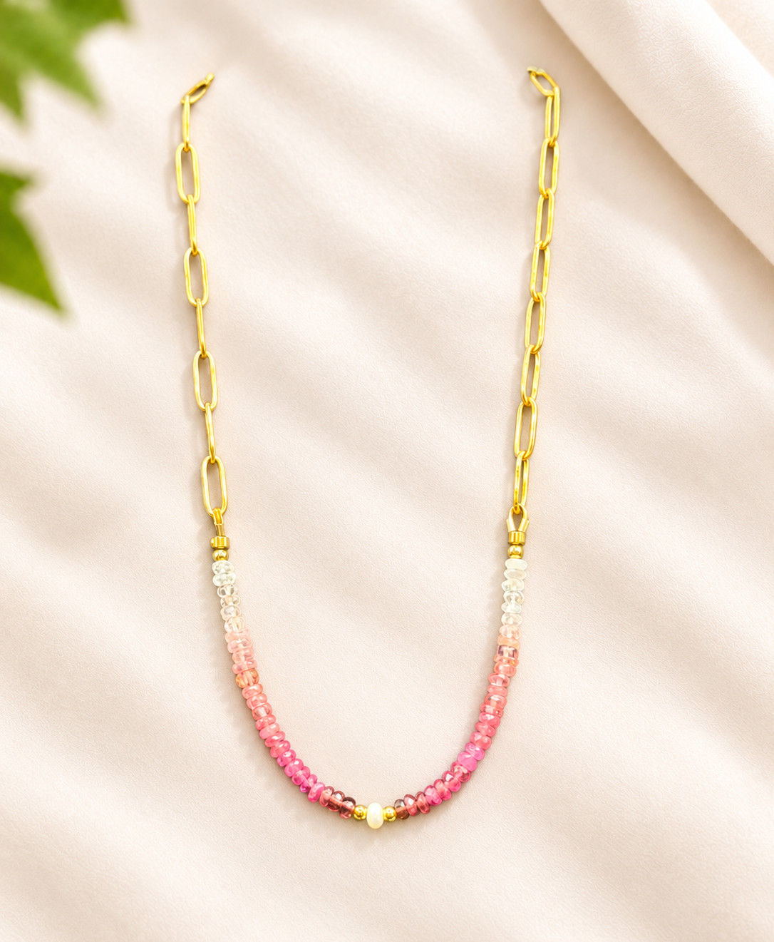 A delicate gold chain necklace with a beaded drop section transitioning from white to pink, displayed on a soft beige fabric background.