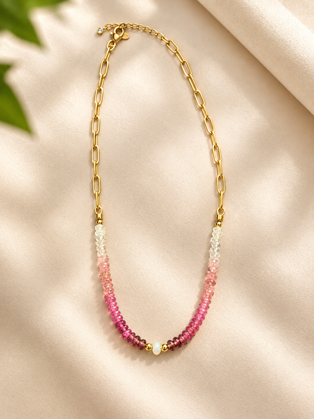 A delicate gold chain necklace featuring two strands of faceted beads that transition from clear at the top to light pink, medium pink, and deep magenta at the bottom, accented by small gold beads, displayed on a cream satin fabric background.