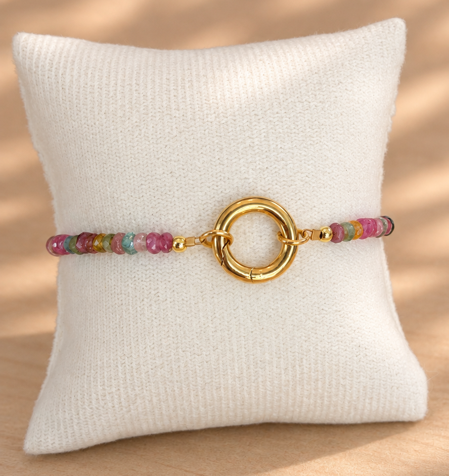 A beaded bracelet with a gold ring clasp displayed on a white textured pillow over a light wood surface with soft shadow patterns.