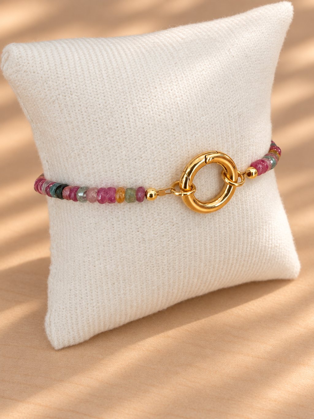 A beaded bracelet with a gold ring clasp displayed on a cream-colored textured pillow, set against a warm beige background with soft shadows.