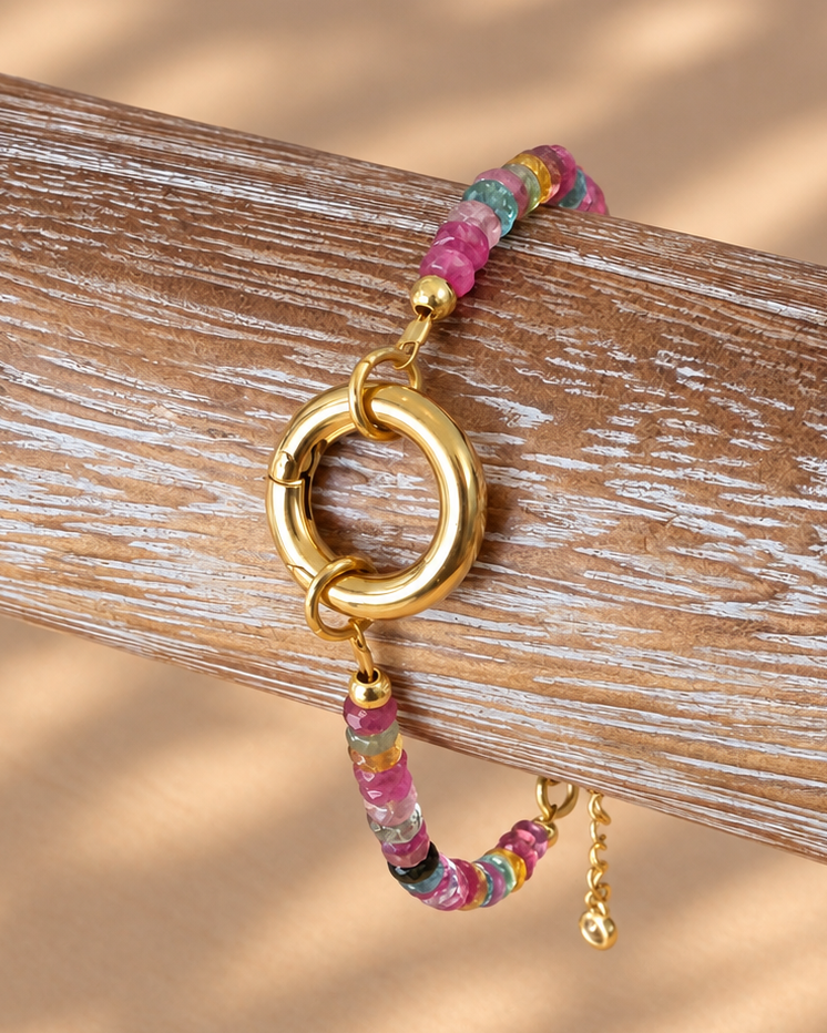 A gold-tone ring clasp bracelet with a colorful floral-patterned cord, displayed on a weathered wooden branch against a warm beige background with soft leaf shadows.