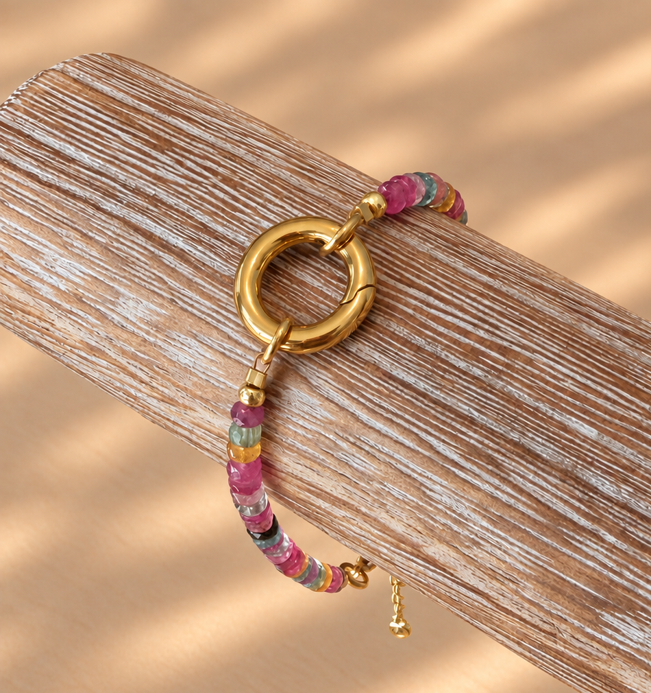 A gold-tone ring charm bracelet with a colorful patterned fabric strap, resting on a textured wooden block against a warm beige background with soft shadow patterns.