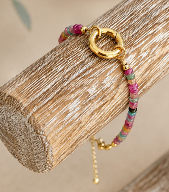 A delicate beaded bracelet with pink, green, and gold beads, featuring a gold heart-shaped clasp and a small dangling gold charm, displayed on a rustic wooden stand.